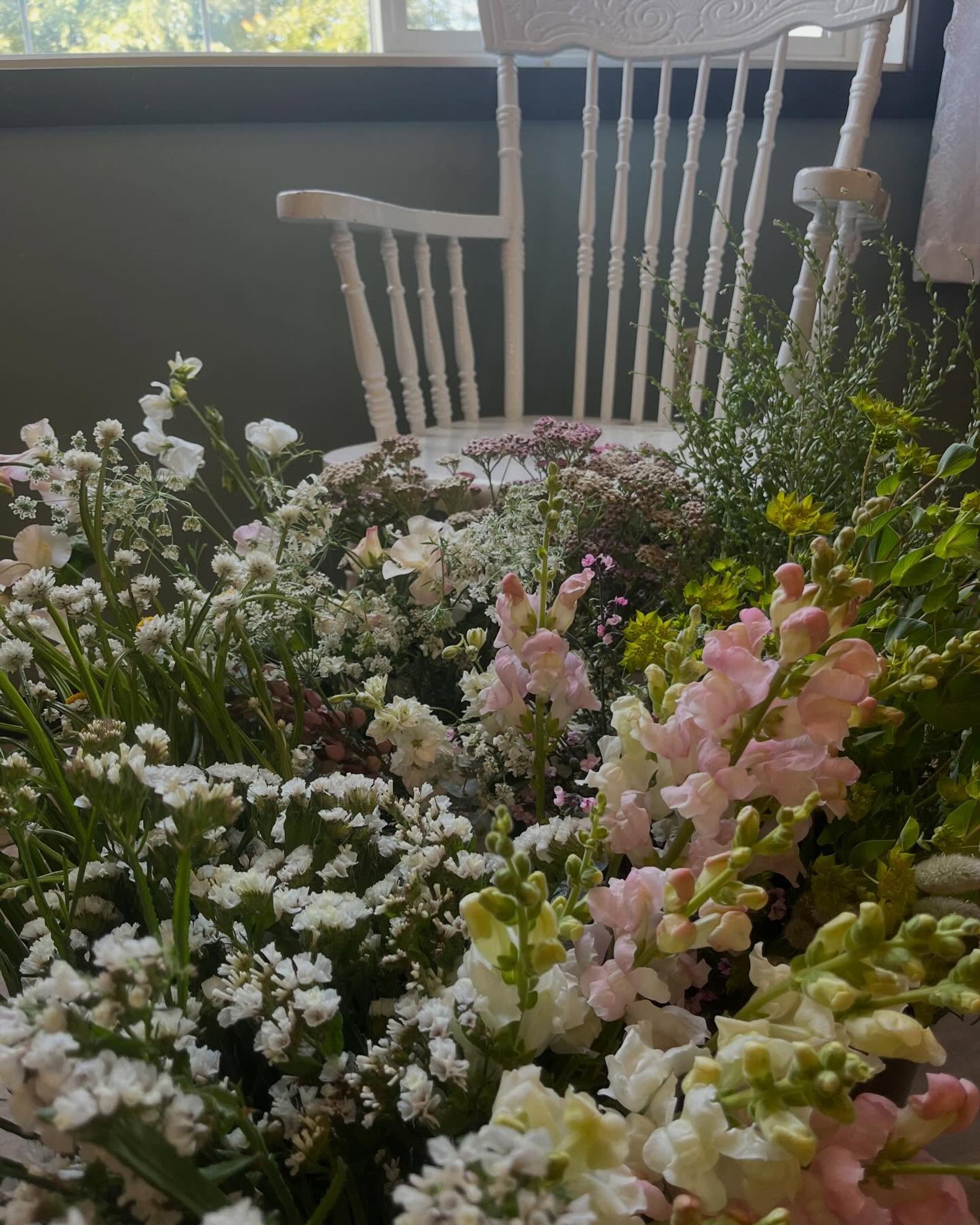 White wooden chair behind dense arrangement of pastel and white cut flowers indoors.