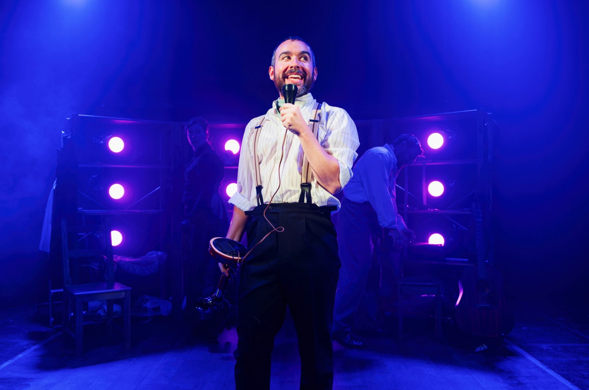 Performer in suspenders holds microphone on stage with purple spotlights and crew.