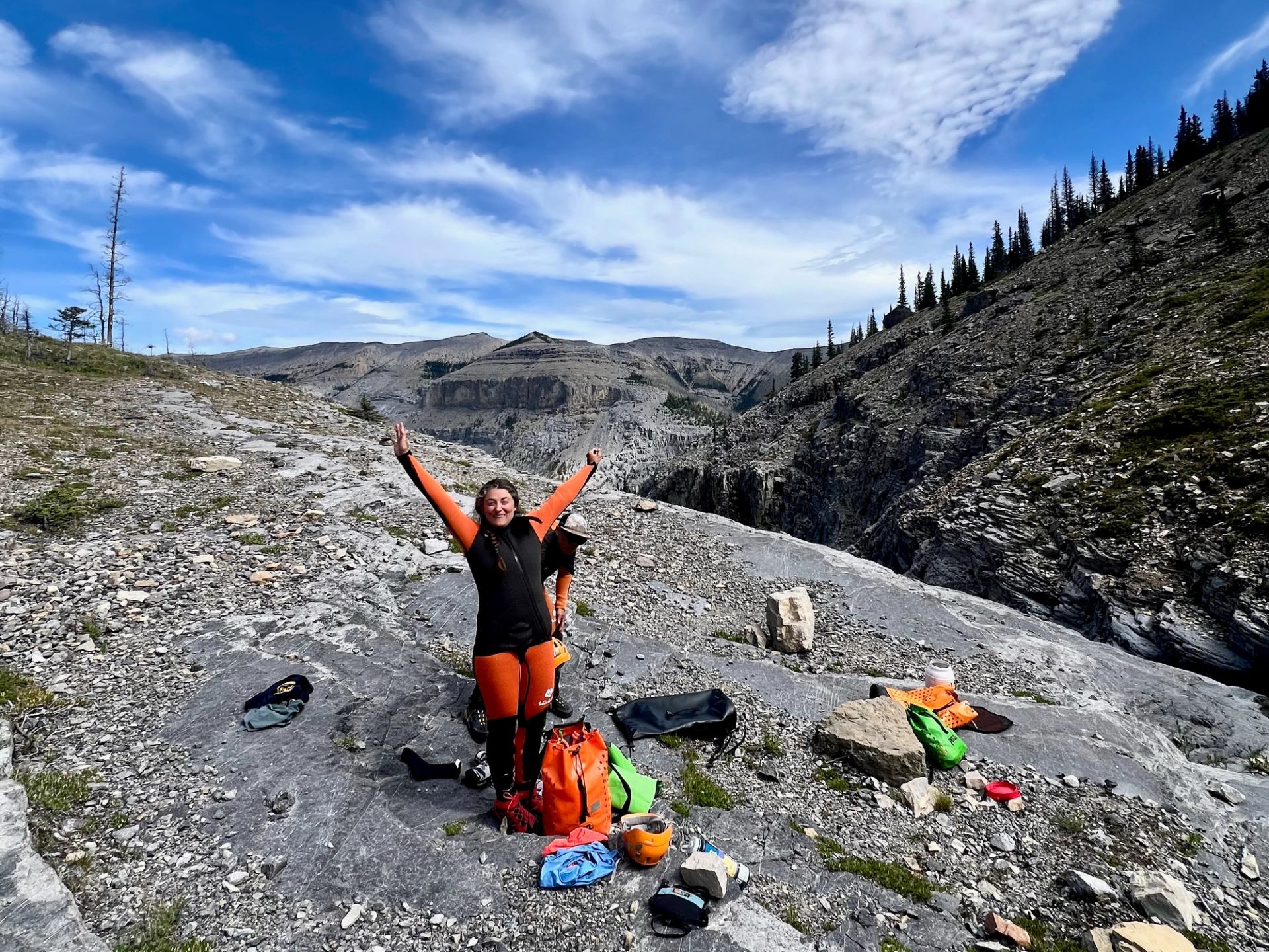 Person standing on rocky terrain with arms raised, gear scattered around.
