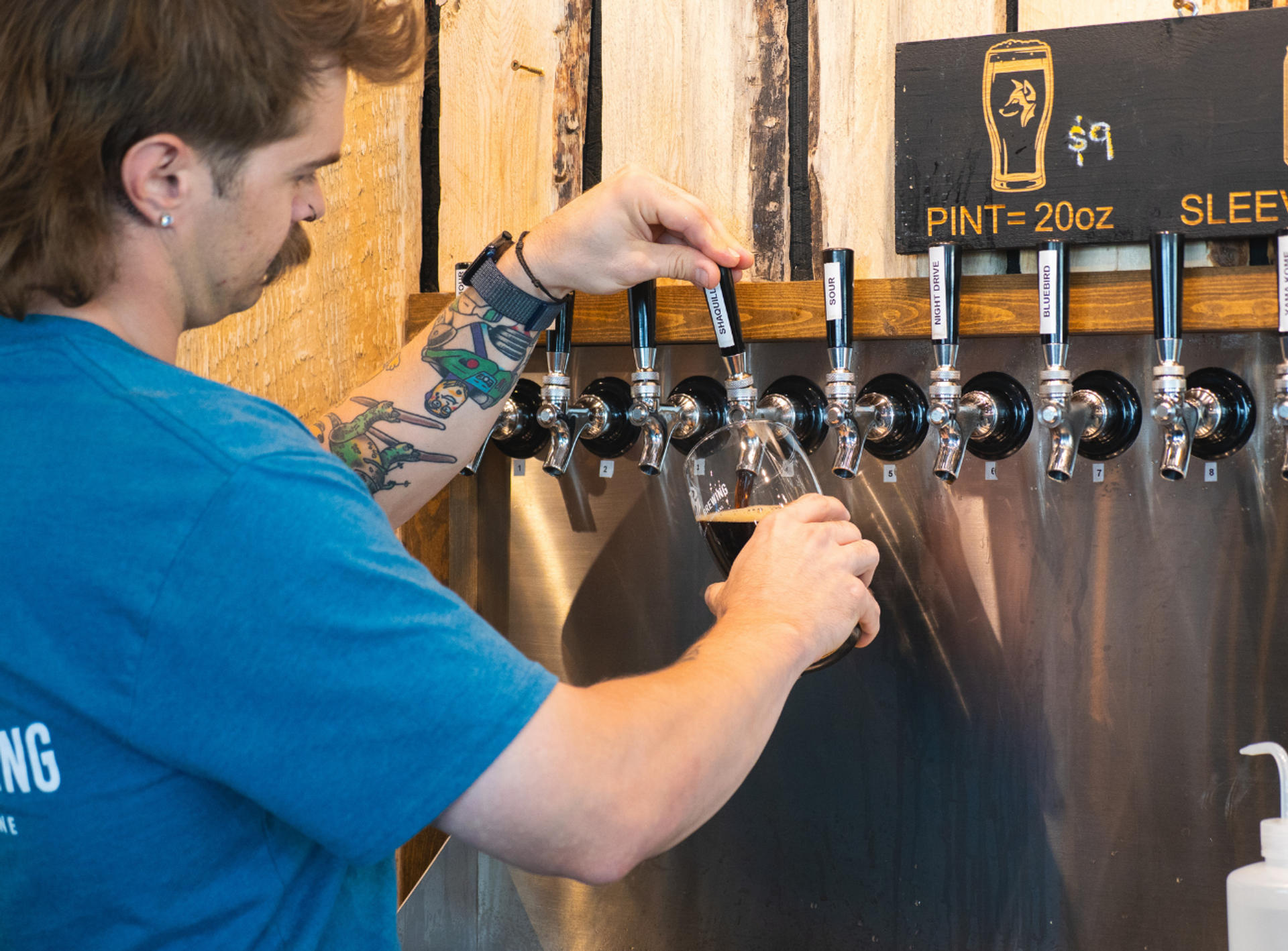 Tattooed bartender pours dark beer into a glass from a row of taps.