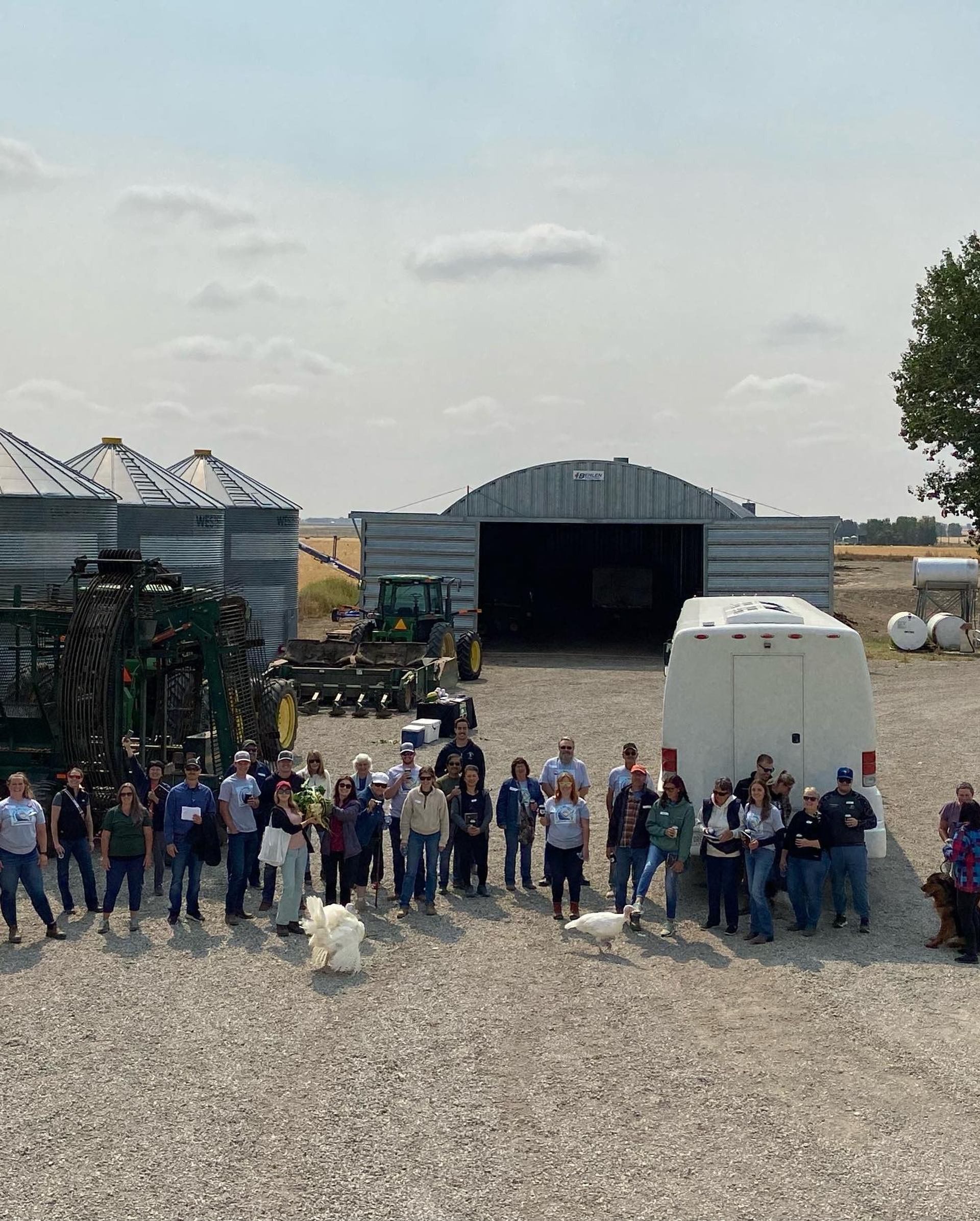 Group of people standing on farmyard with tractors, grain bins, bus and turkeys.