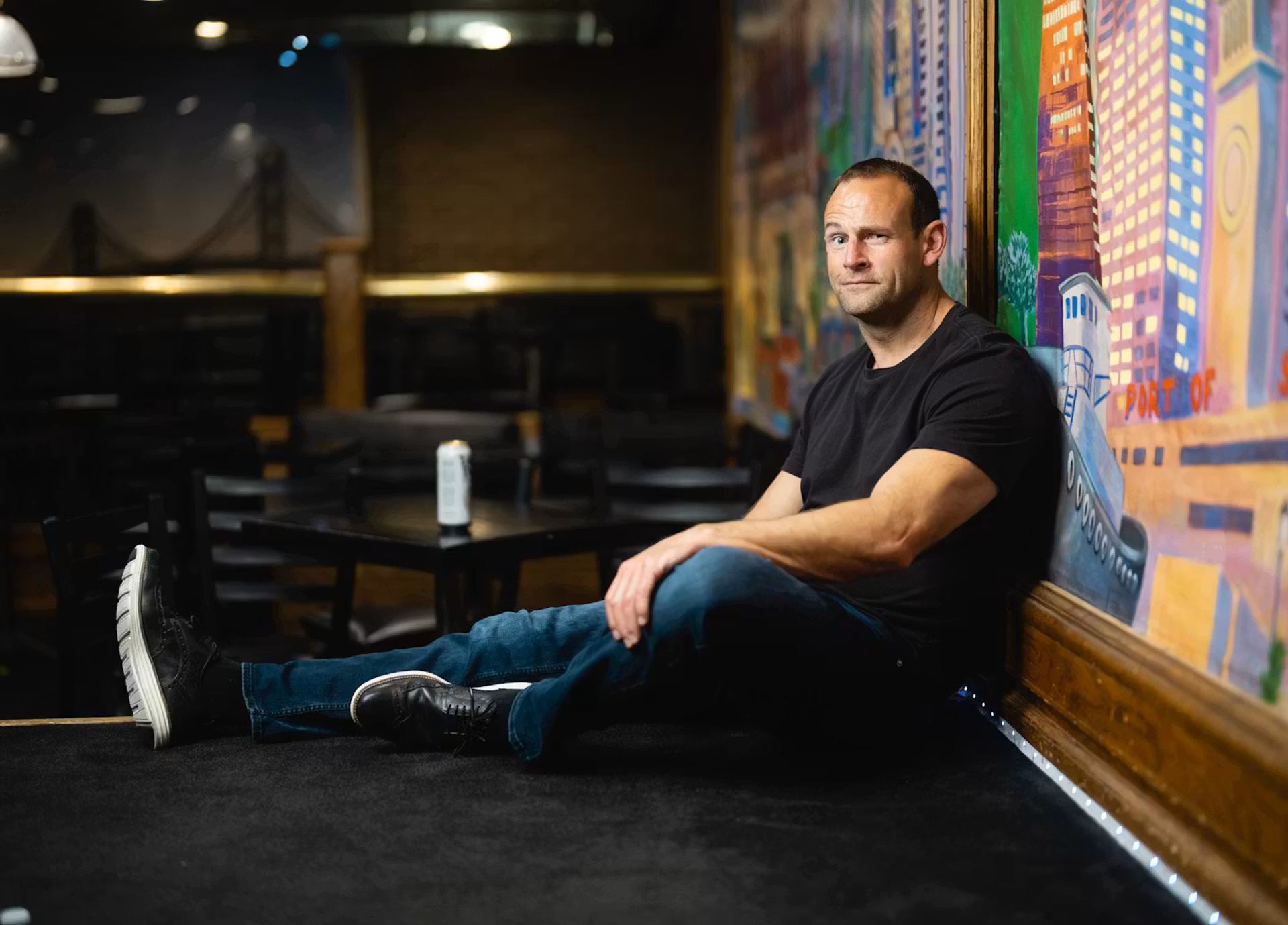 Man in black T-shirt sits on stage floor beside colourful mural in dim venue.