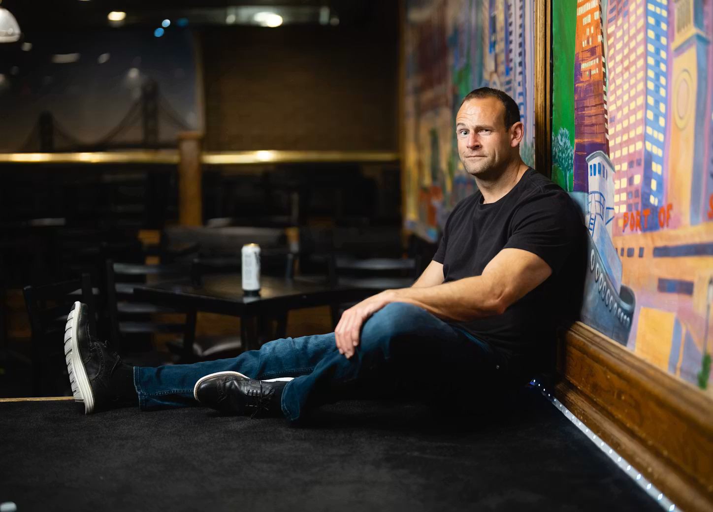 Man in black T-shirt sits on stage floor beside colourful mural in dim venue.