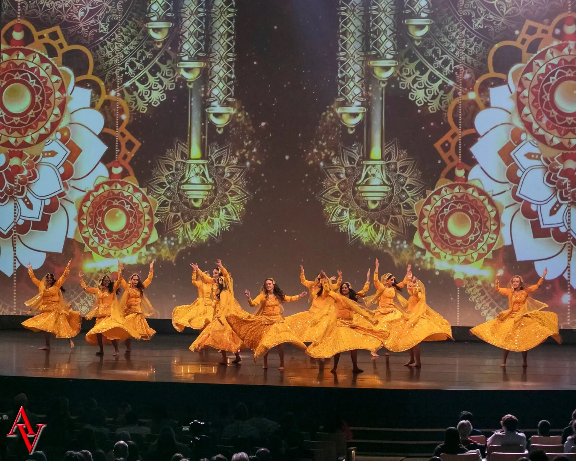 A group of dancers dressed in sparkling yellow-gold costumes performing synchronized spins on stage, with a colorful mandala and ornamental pattern projected in the background.
