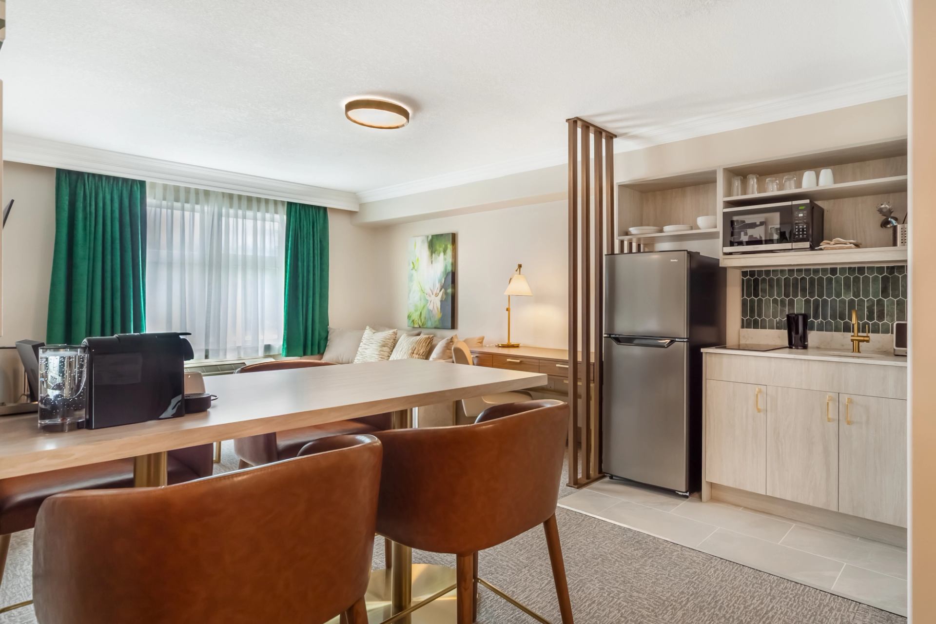 Modern hotel suite with a dining table, leather chairs, kitchenette featuring a fridge, microwave, and coffee maker.