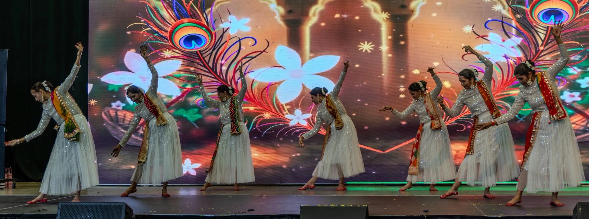 Dancers in white dresses perform before a colourful floral and peacock feather backdrop.