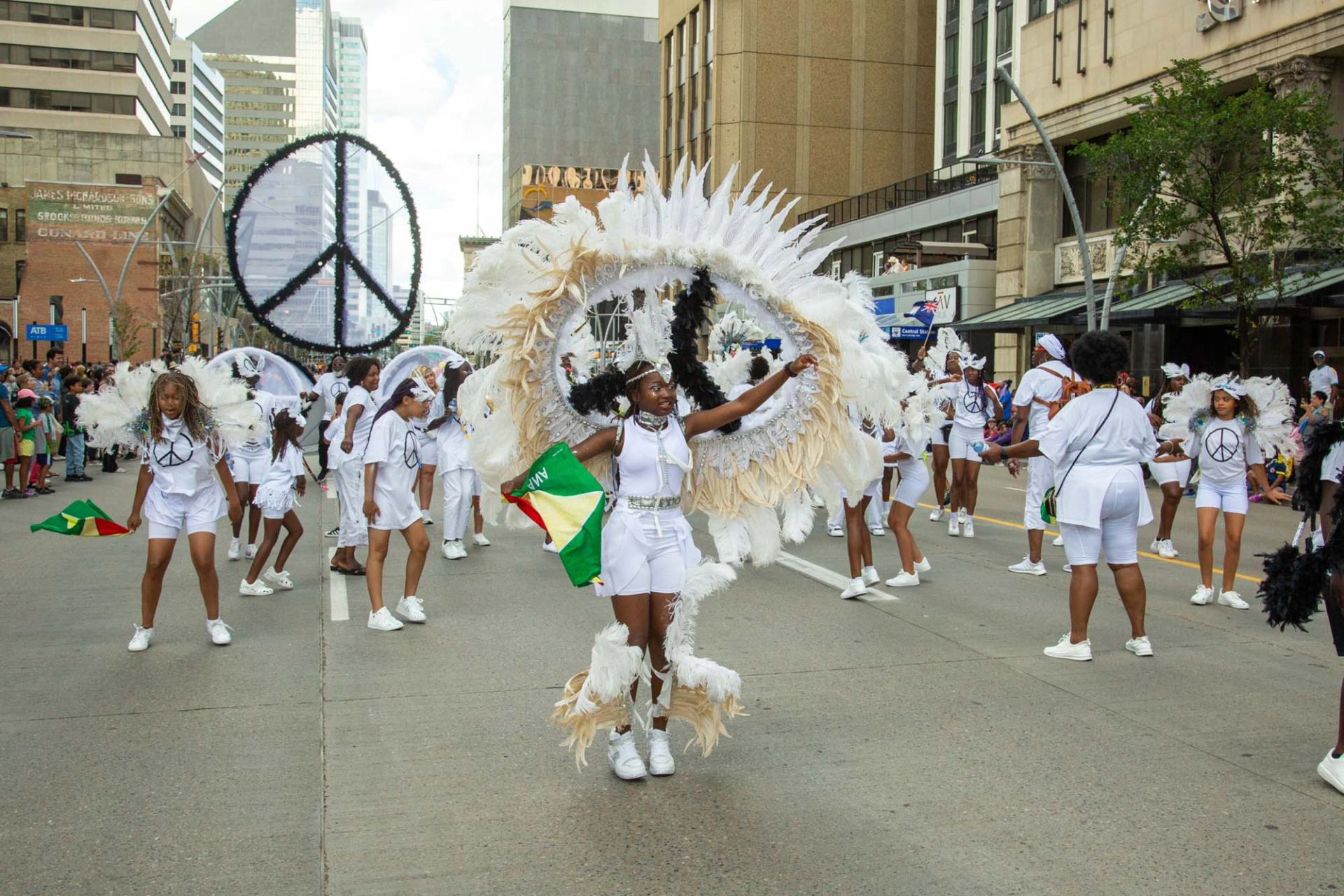 Parade dancers in white costumes with feathered wings and peace symbols on city street.