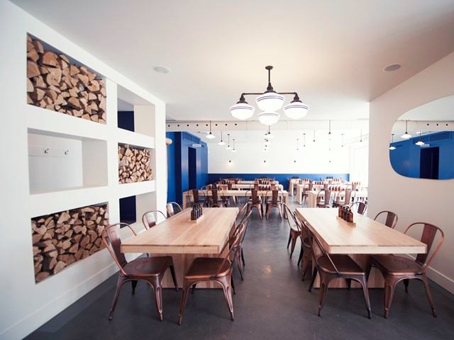 Modern dining room with wood tables, metal chairs, firewood shelves, and chandelier lighting.