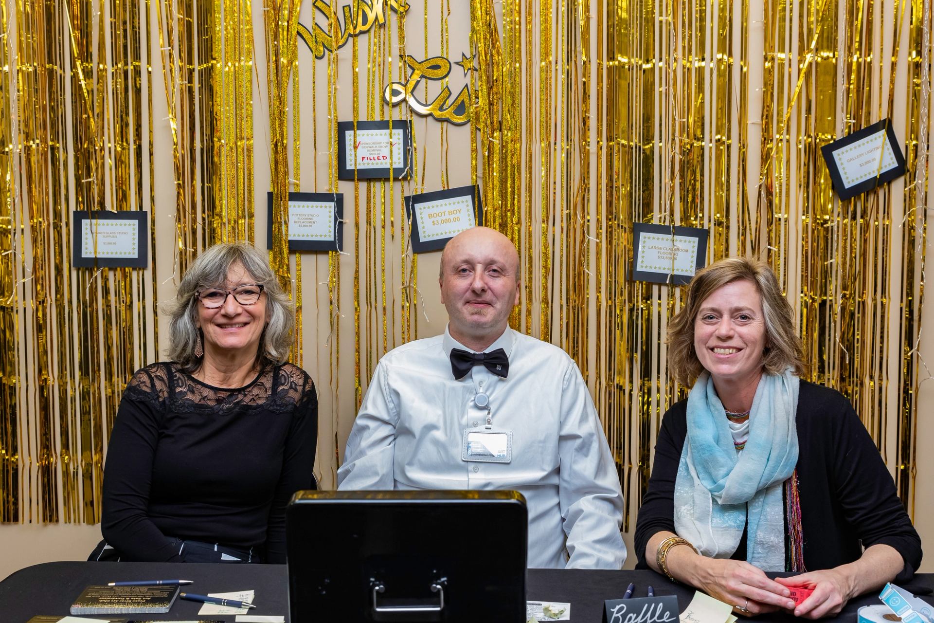 Three people seated at table with gold tinsel backdrop and “Wish List” sign.