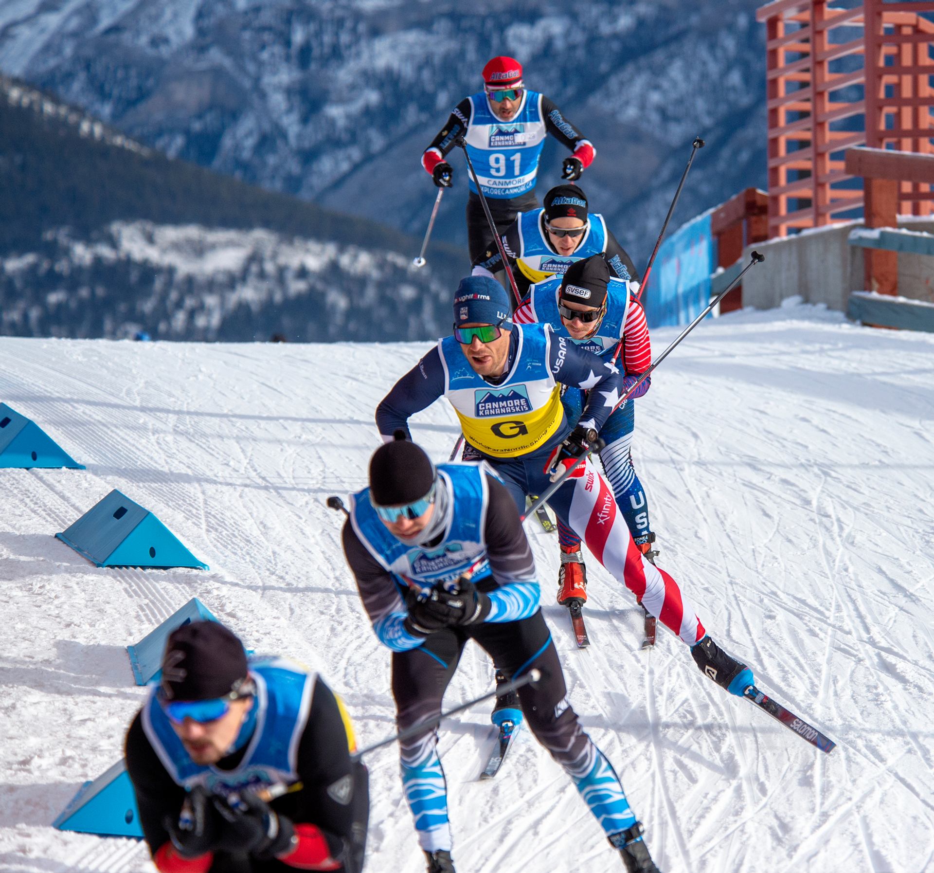 Para cross-country skiers race downhill with snowy mountains and bright blue skies behind.