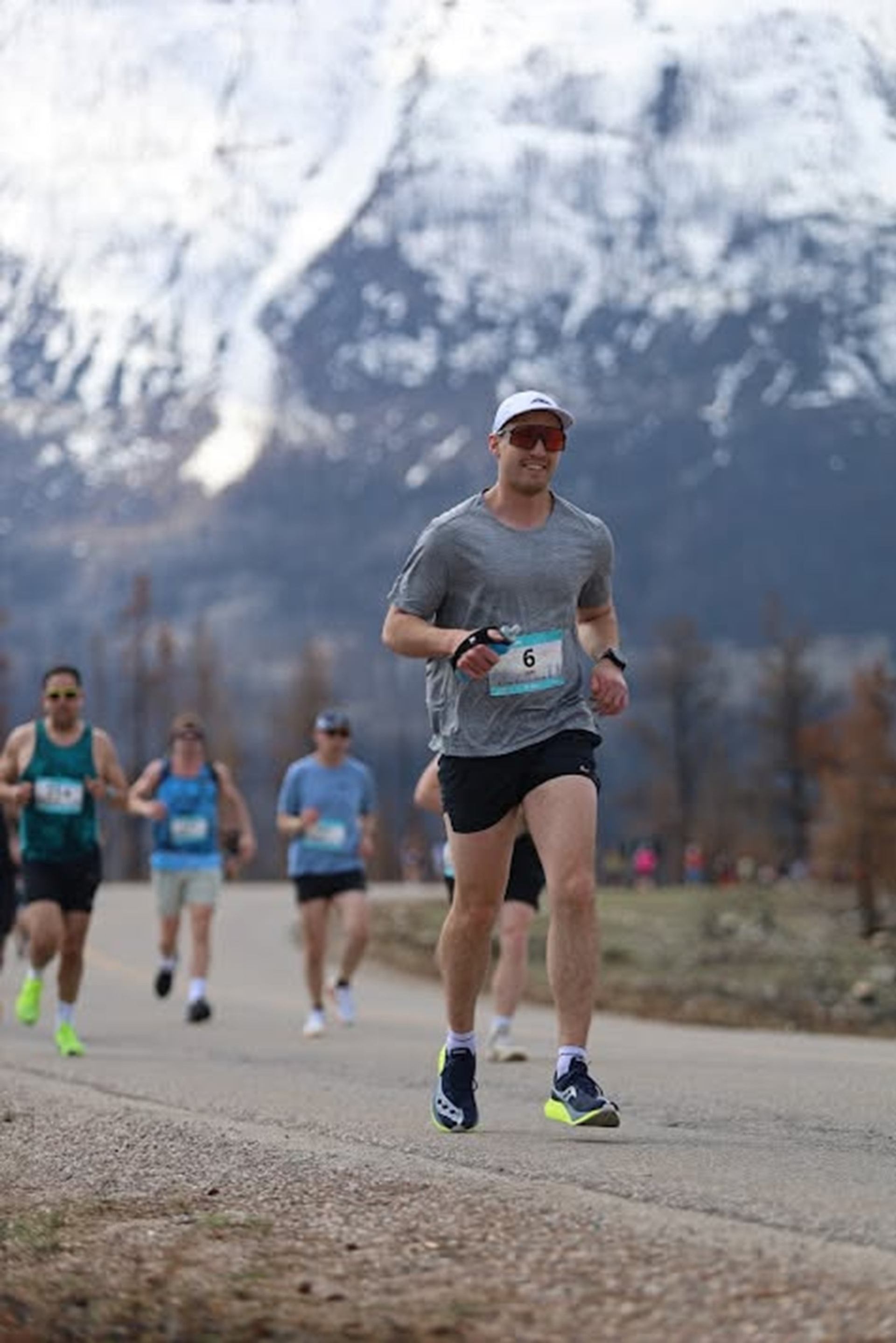 Road runners wearing race bibs move along paved path below snow-covered mountain.