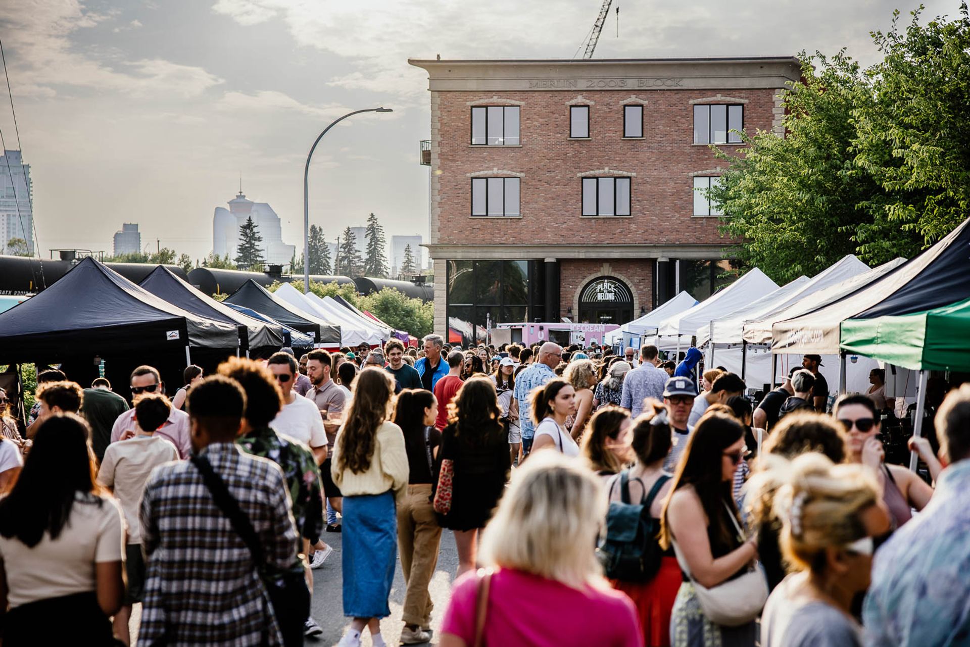 Crowd walks between vendor tents at outdoor market with brick building and city skyline.