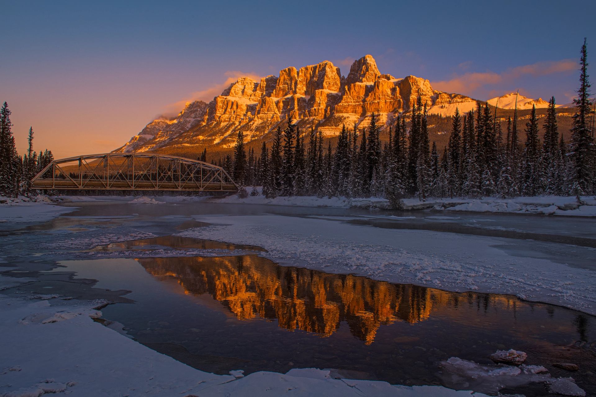 Snowy riverbank with frozen water reflecting sunlit mountains and a bridge at sunset.