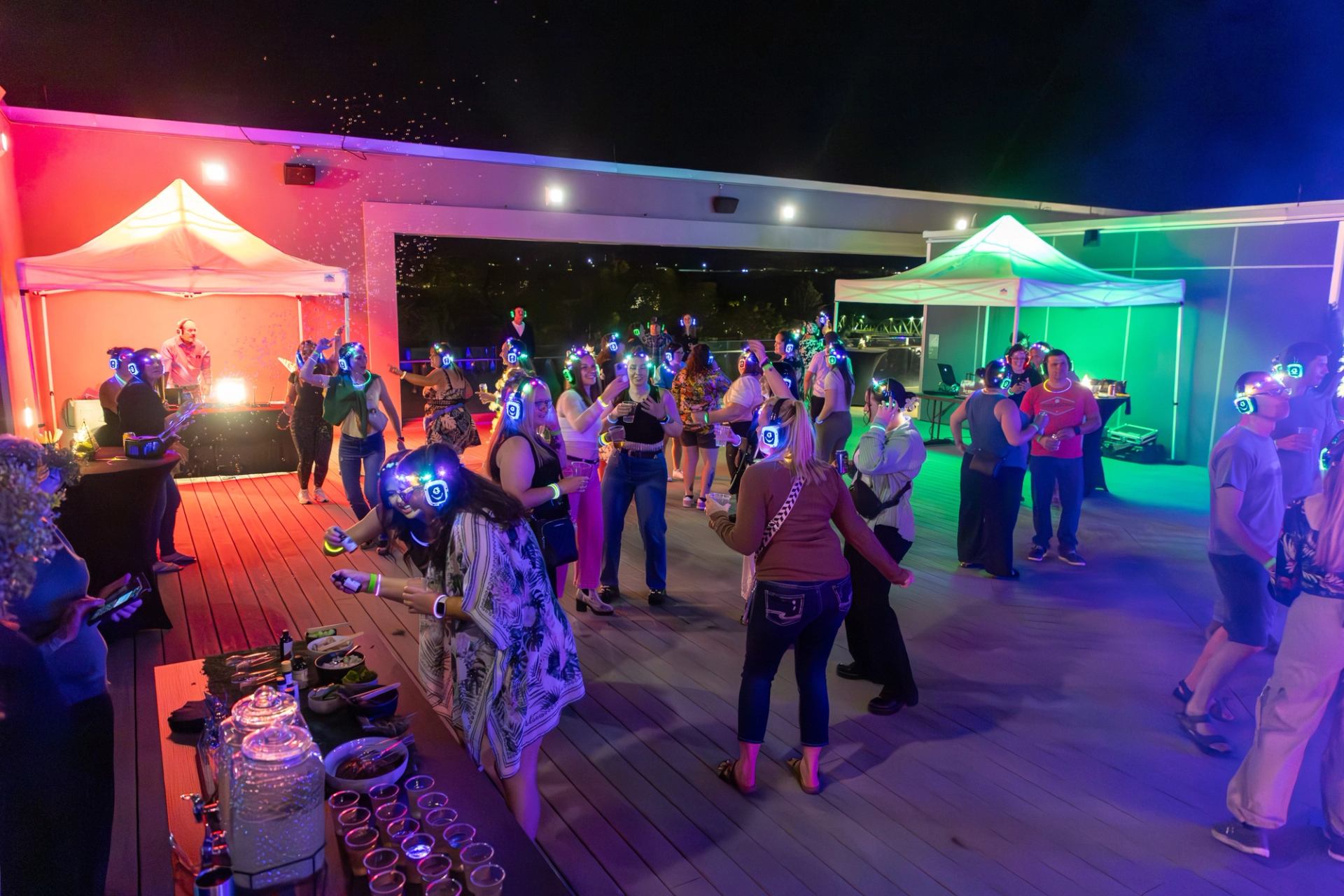 Crowd wearing glowing headphones dances on rooftop deck with colourful lights and tents.