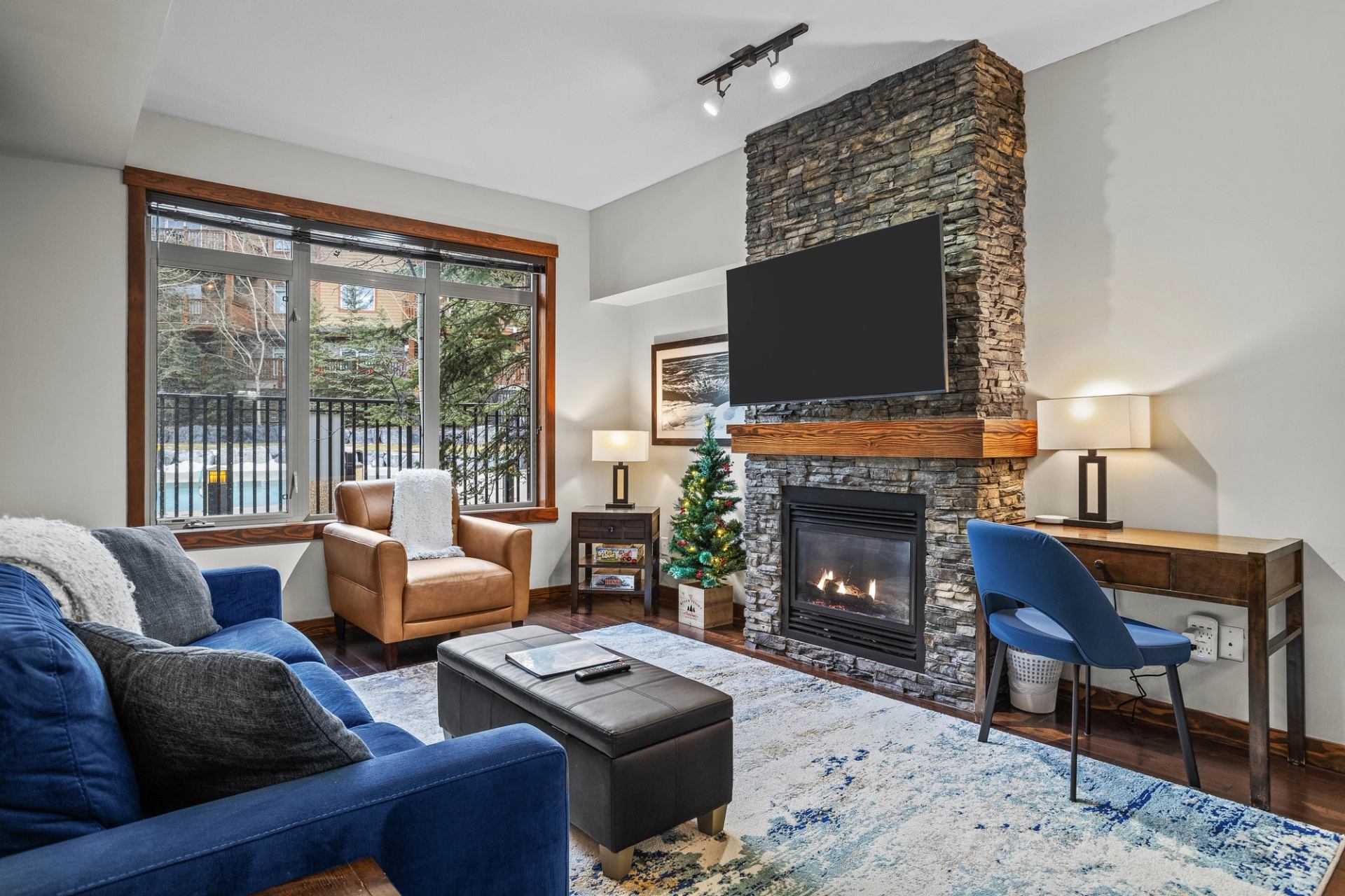Living room with stone fireplace, wall-mounted TV, blue sofa, desk and window view.