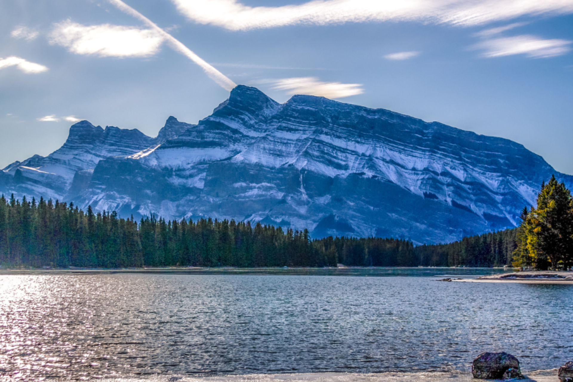 Snowy mountain rising above a lake with a forested shoreline on a bright day.