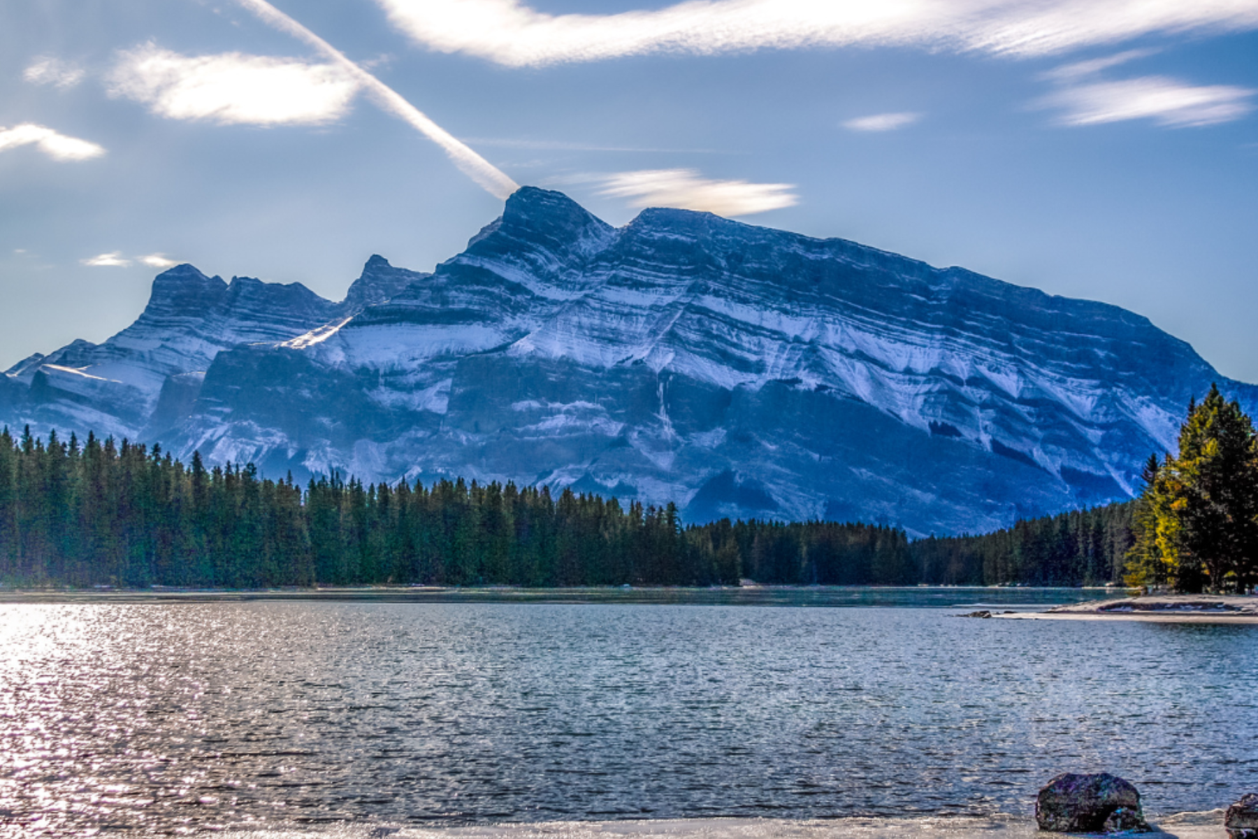 Snowy mountain rising above a lake with a forested shoreline on a bright day.