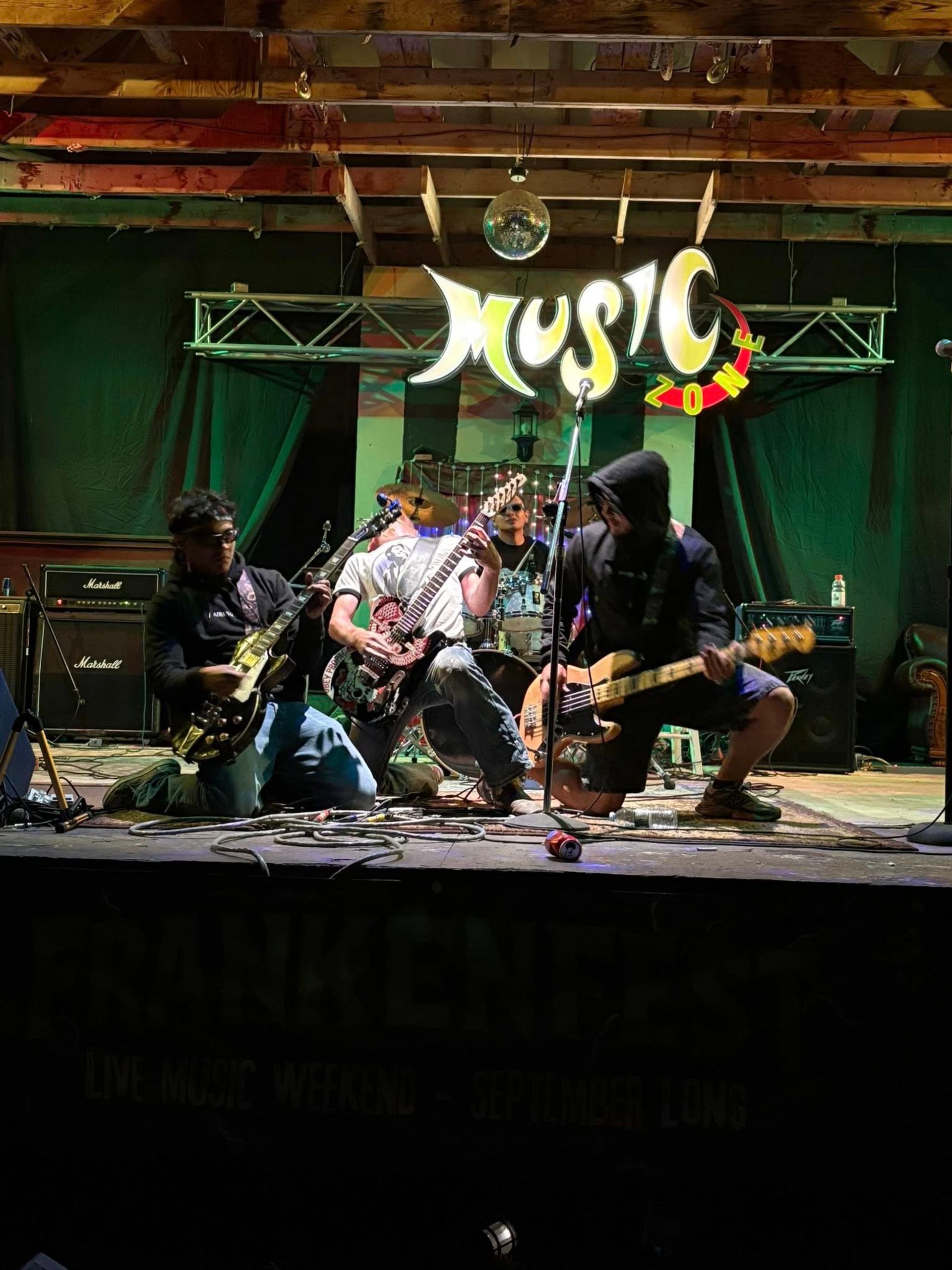Three musicians kneel on stage, playing electric guitars and bass in front of a "MUSIC ZONE" sign.