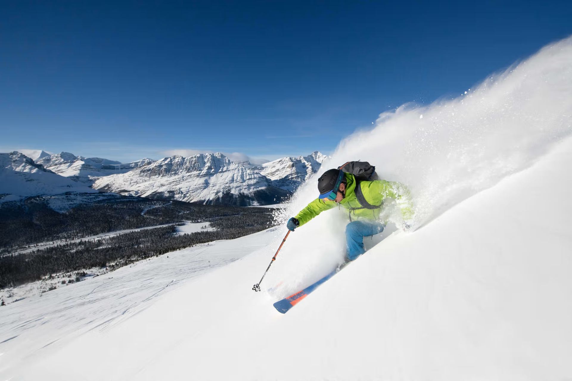 A skier in a green jacket makes a turn on a snowy mountain, kicking up powder, with distant peaks under a clear blue sky.