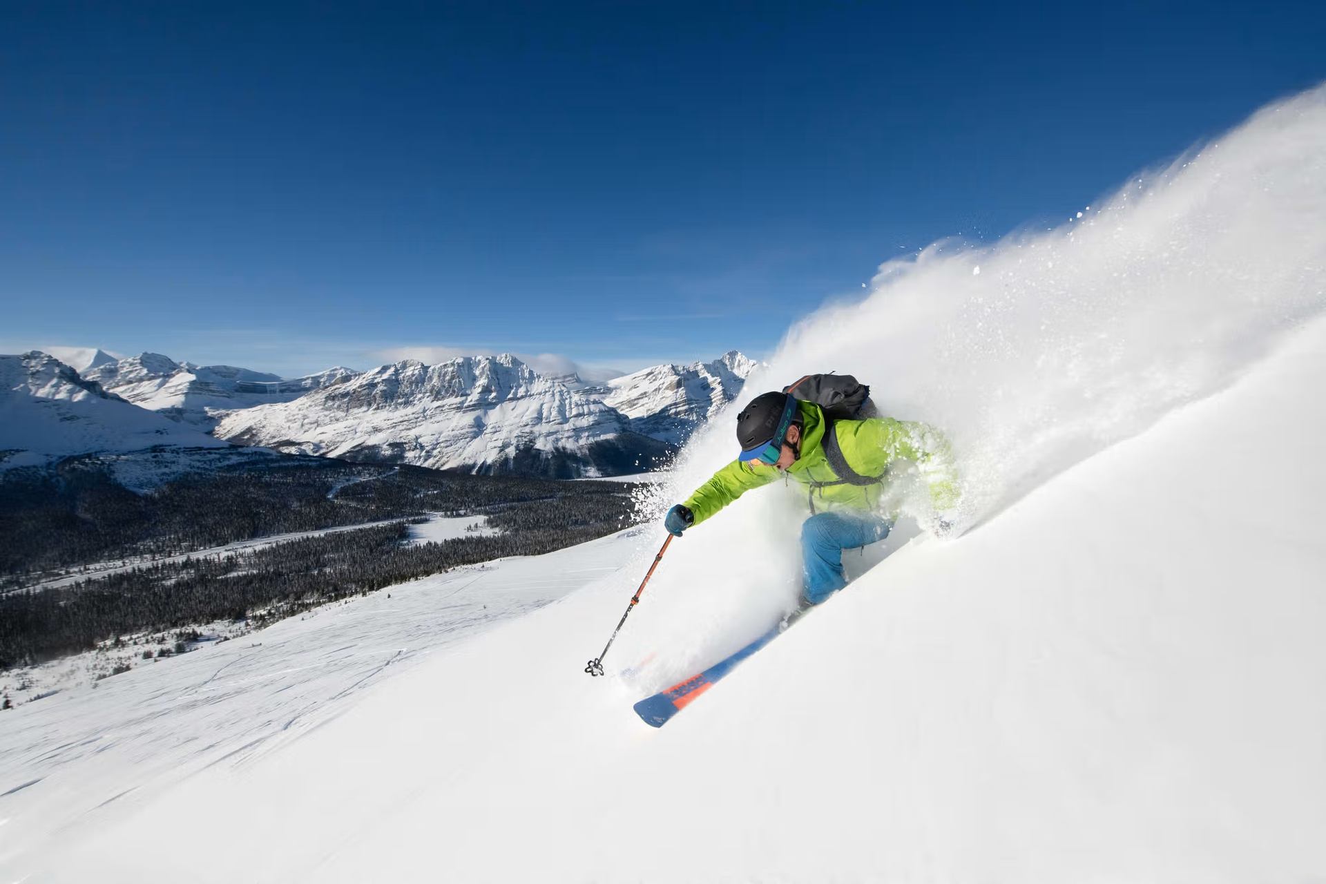 A skier in a green jacket makes a turn on a snowy mountain, kicking up powder, with distant peaks under a clear blue sky.