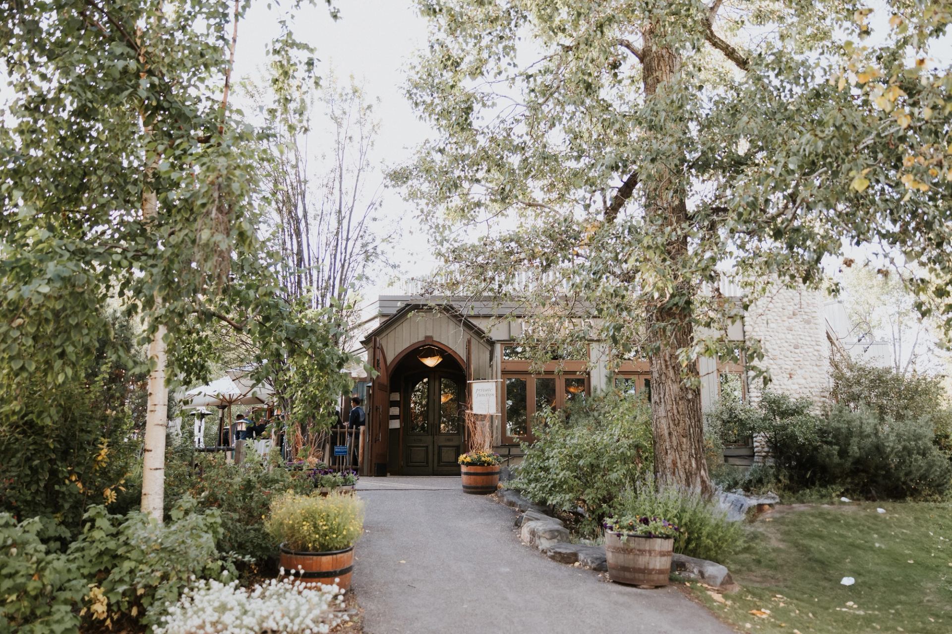 Tree-lined path leads to a rustic building entrance surrounded by gardens and flower planters.