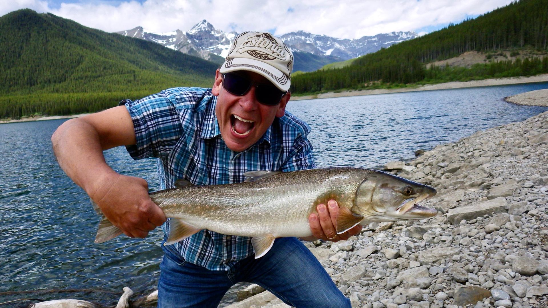 Angler in plaid shirt holds large fish beside rocky lakeshore with mountain backdrop.