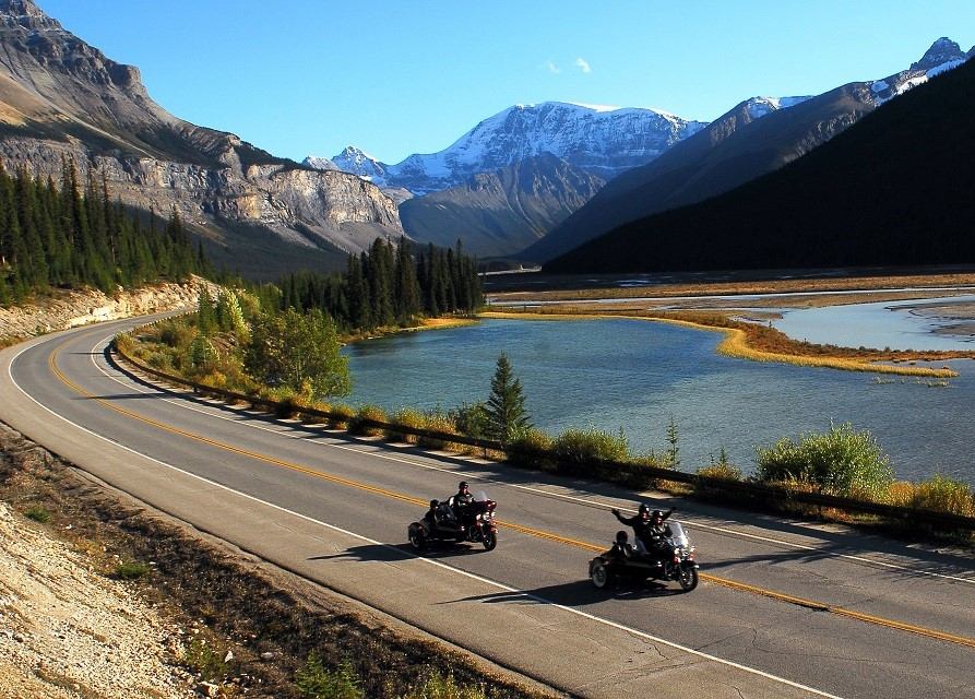 Two motorcyclists ride along a winding road by a tranquil lake, surrounded by majestic snow-capped mountains under a clear blue sky.