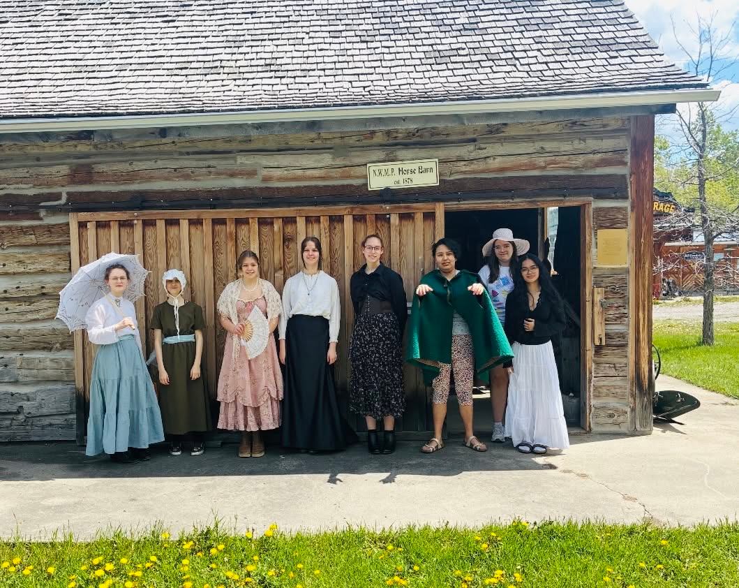 Group of people in varied historical costumes standing outside wooden NWMP Horse Barn.