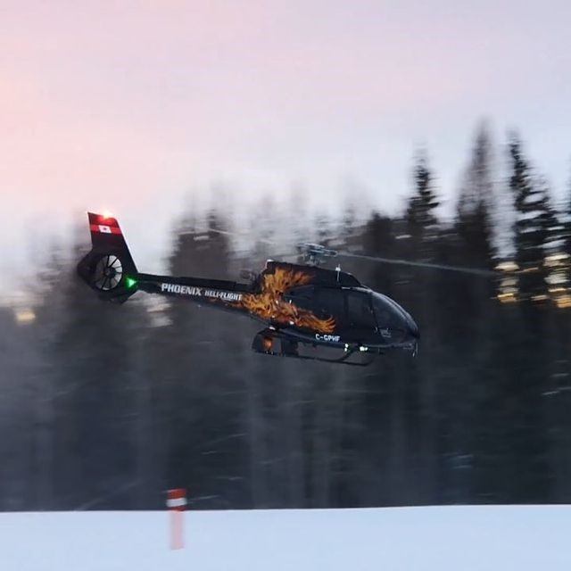 Black and red helicopter flying low over snowy terrain with trees in the background at dusk.