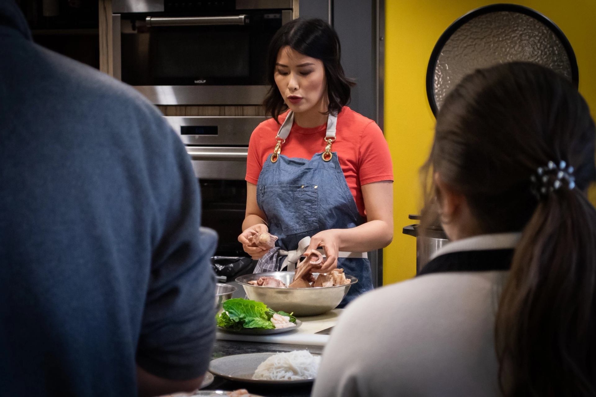 Cook in blue apron slices food at kitchen table during group cooking class.