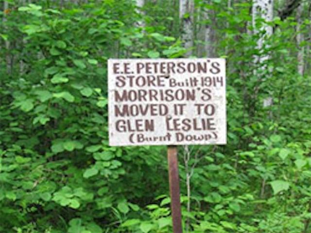 Wooden sign reading “E. E. Peterson’s Store” on a post beside green trees.