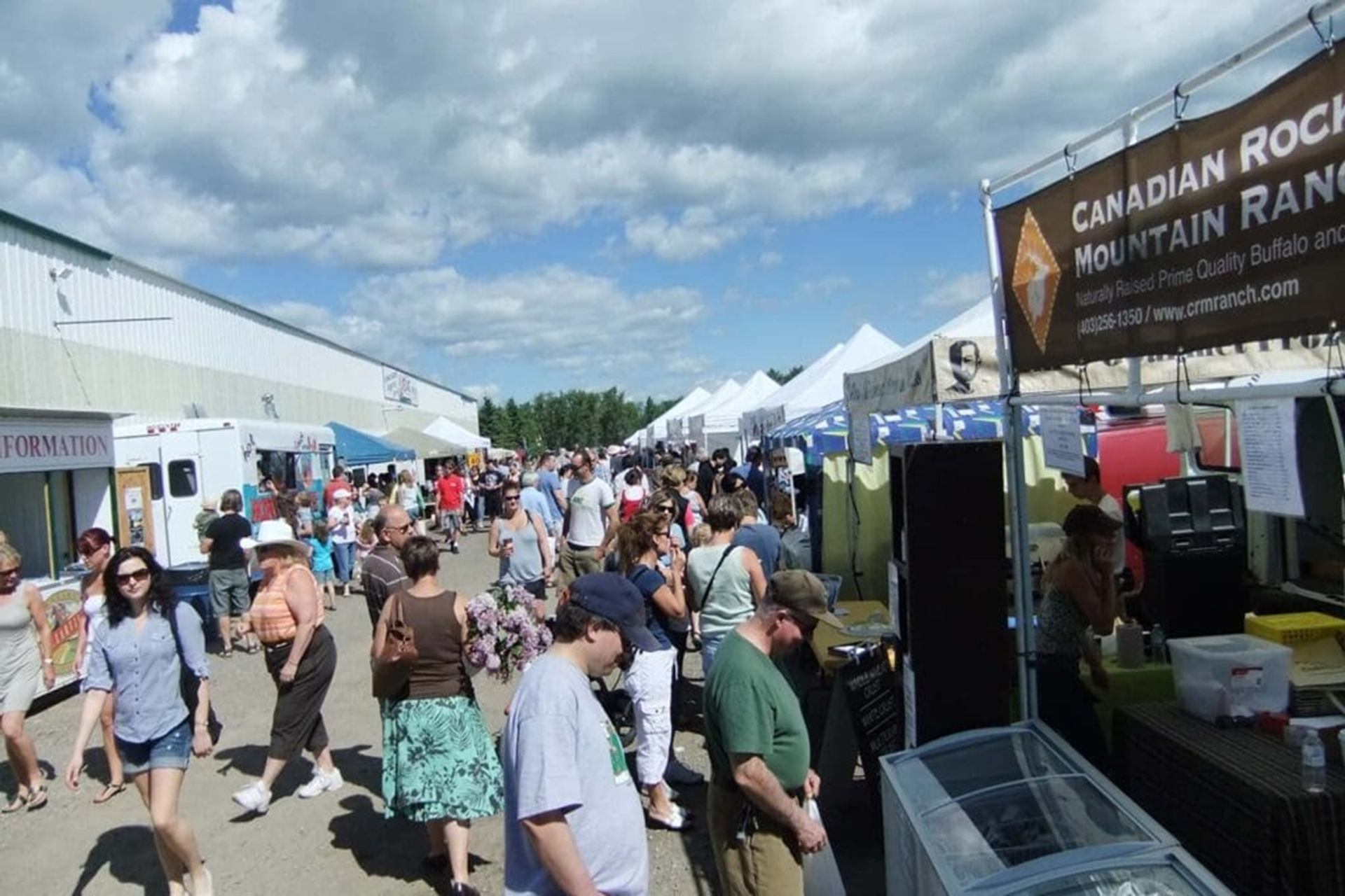 Outdoor market crowd walking between vendor tents and food trucks under partly cloudy sky.