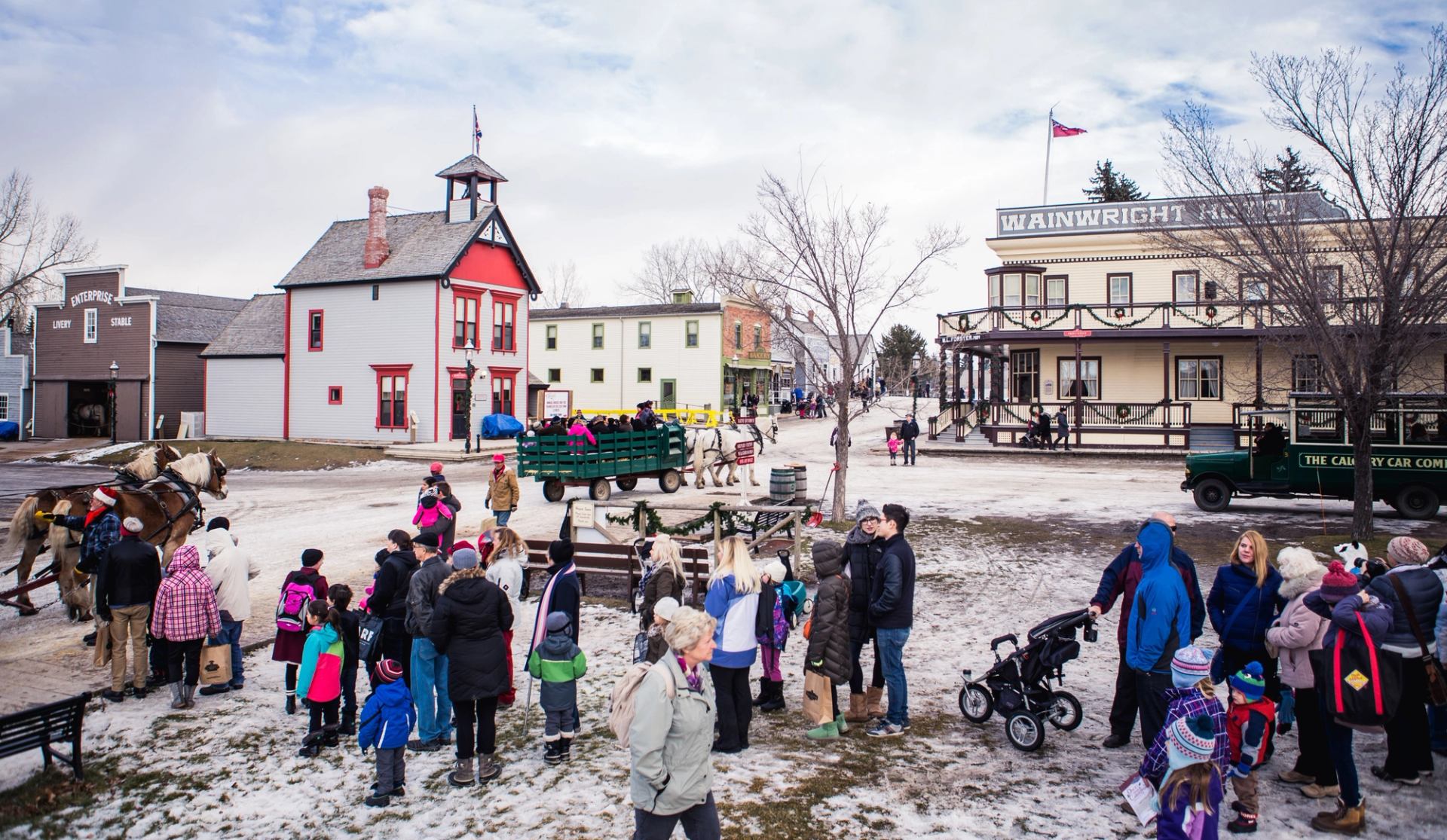 Crowds gather in a snowy village square for festive activities and horse-drawn wagon rides.