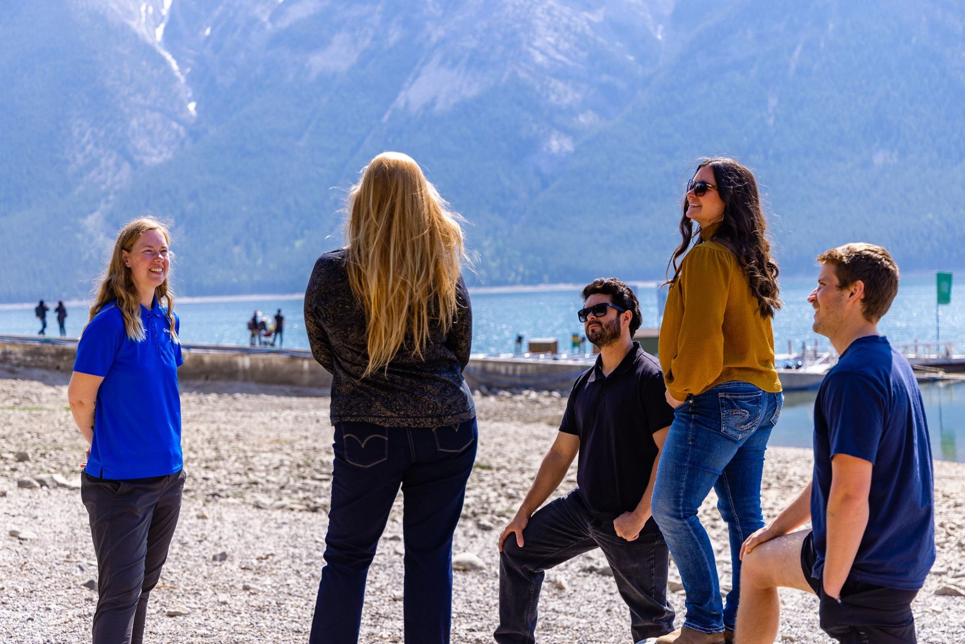 Group of people talking on a rocky lakeshore with mountains and docks behind.