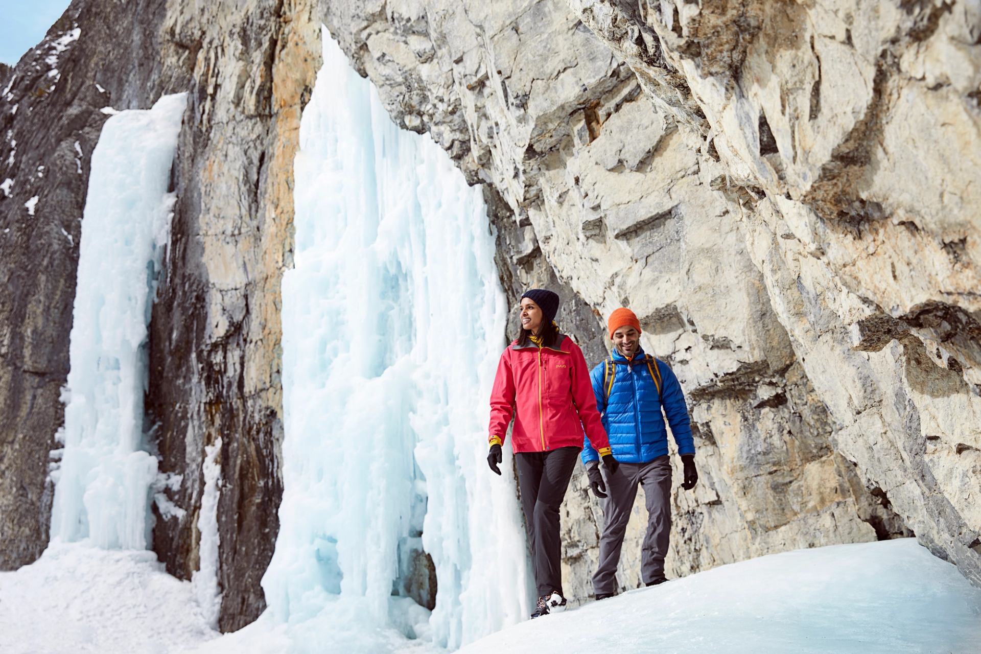 Two hikers in winter gear walk past a towering frozen waterfall.