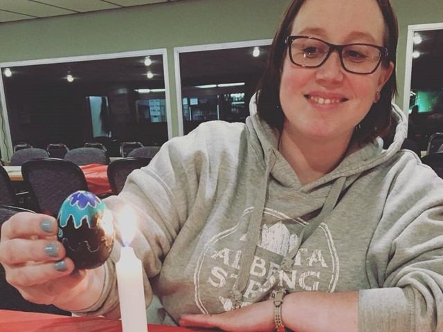 A smiling woman wearing glasses holds a partially decorated Pysanka egg near a lit candle.