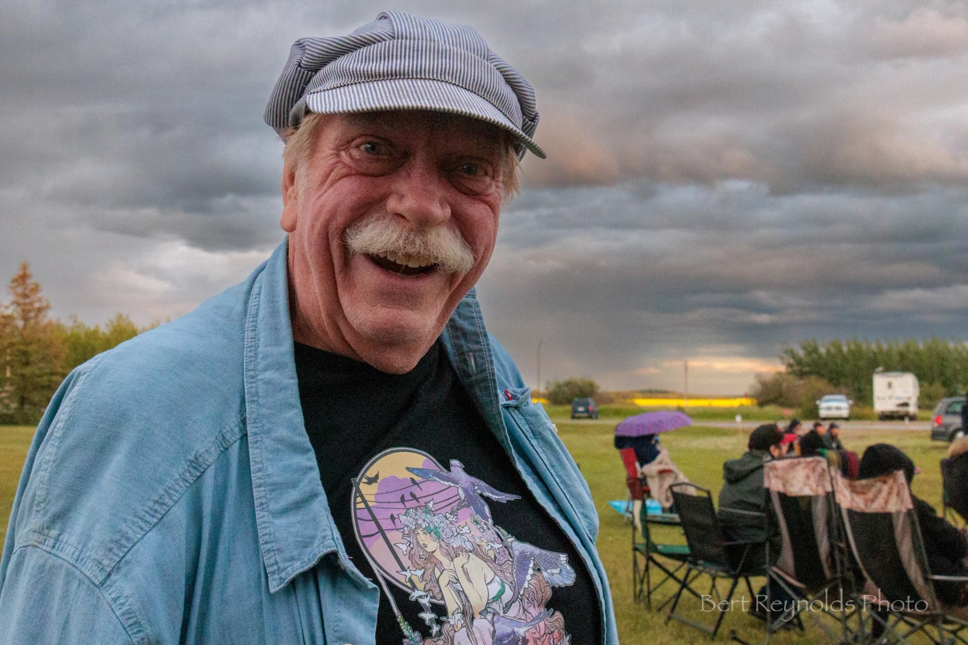 Older person in striped cap at outdoor event, people seated on lawn chairs behind.