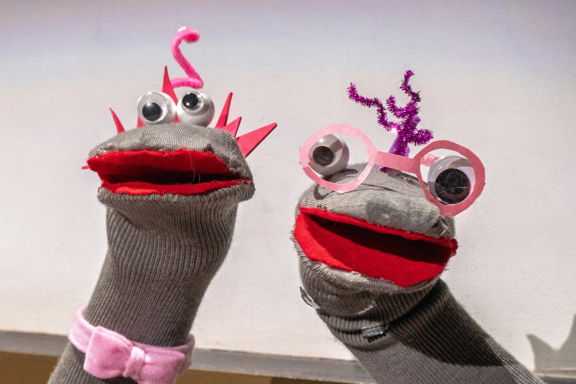 Gloved hands holding puppet faces with red lips and pink circles, purple tinsel on top.