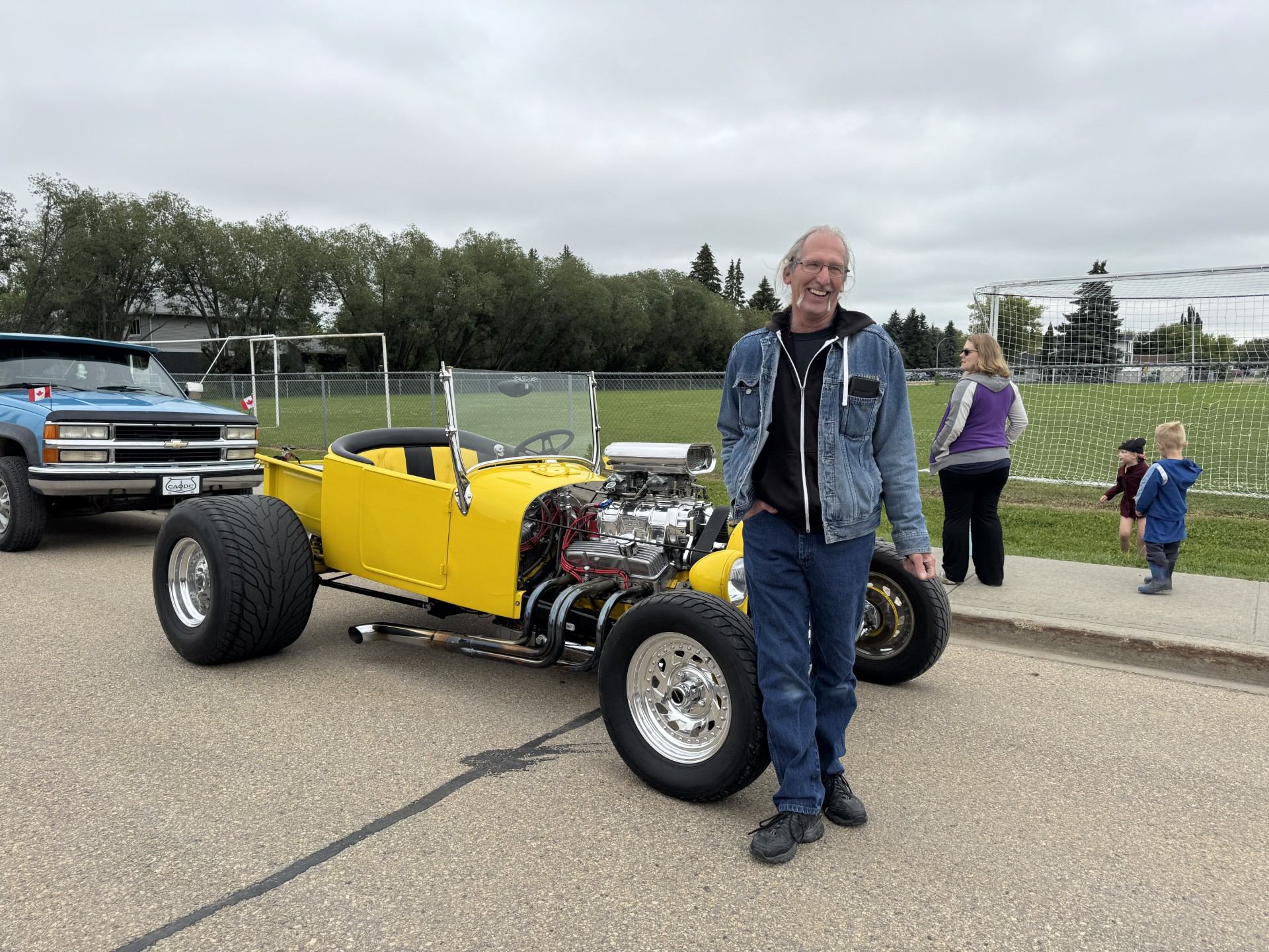 Man standing beside yellow hot rod car near sports field and parked blue truck.