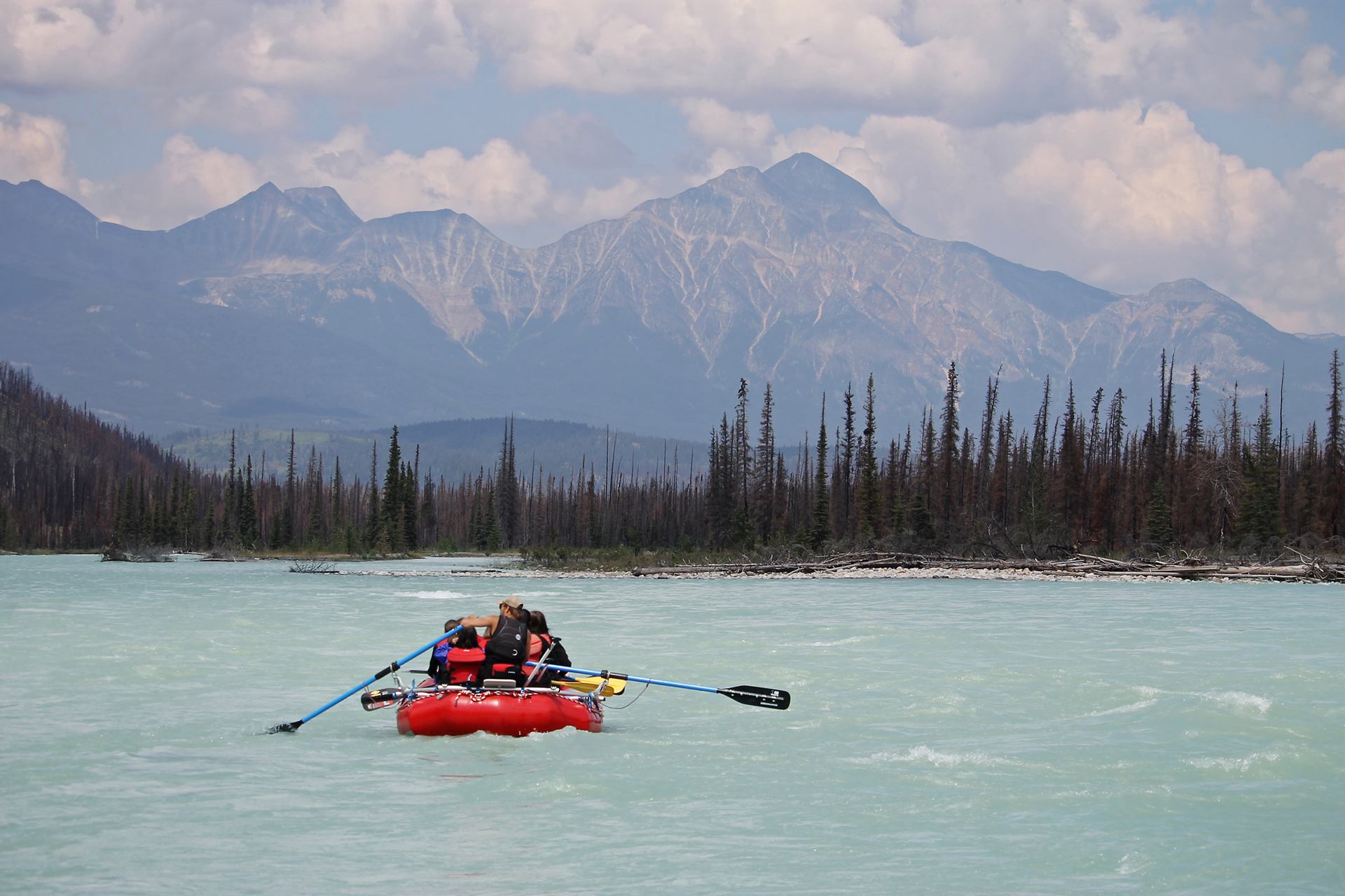 A red raft with people floats on a pale blue river, flanked by dark trees and backed by jagged mountains under a partly cloudy sky.