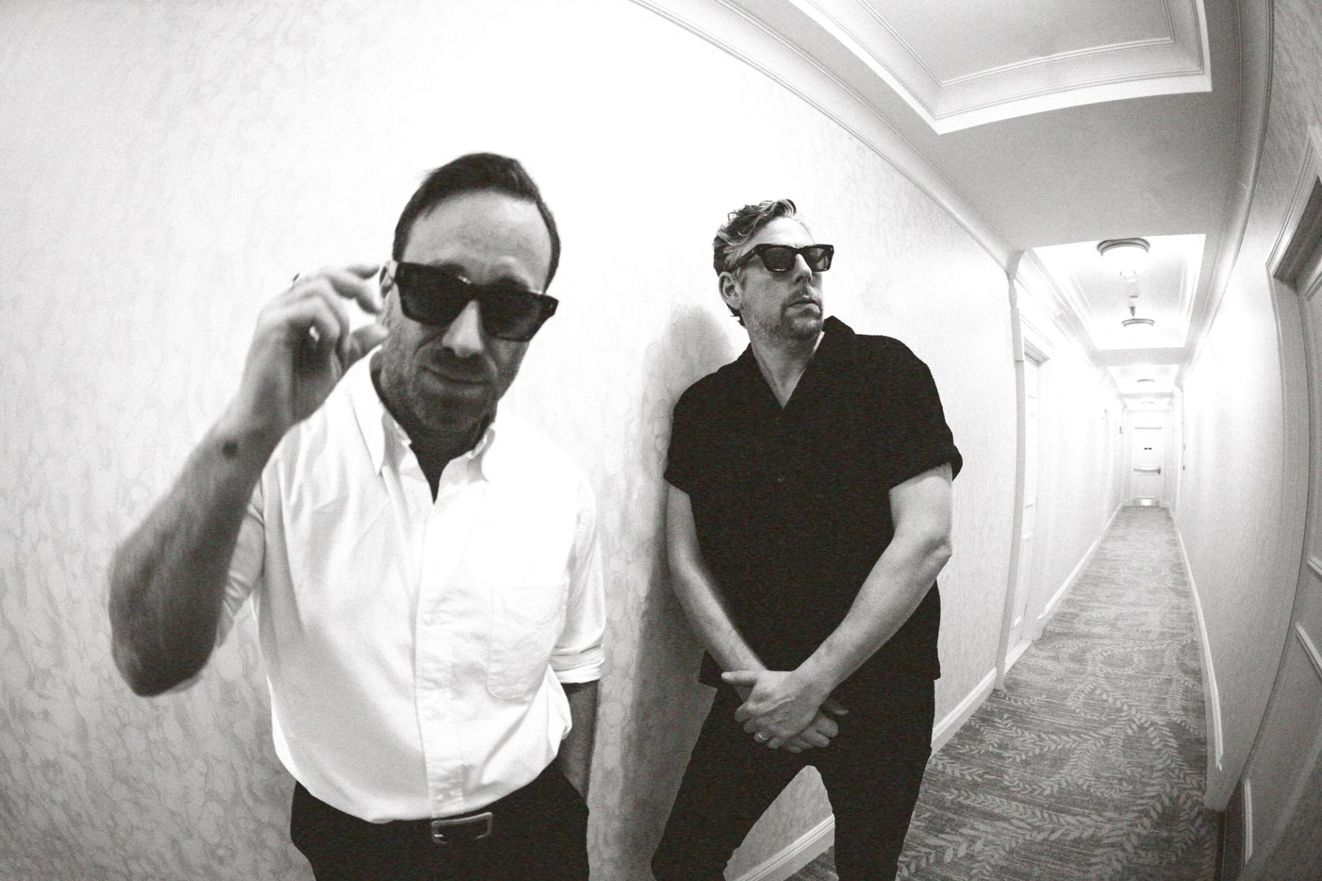 Wide-angle black and white photo of two men in sunglasses in a hotel hallway.