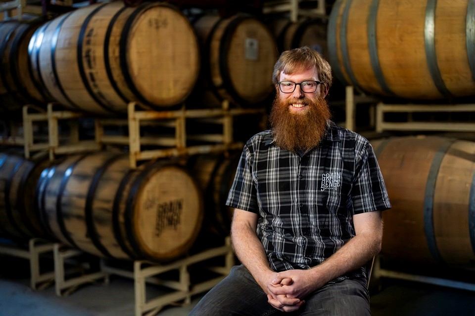 Bearded man in plaid shirt seated in front of stacked wooden barrels at brewery.