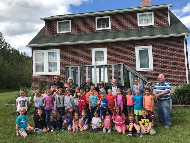 A group of children and adults pose happily in front of a two-story red brick house with a green roof and white trim. The sky is partly cloudy.