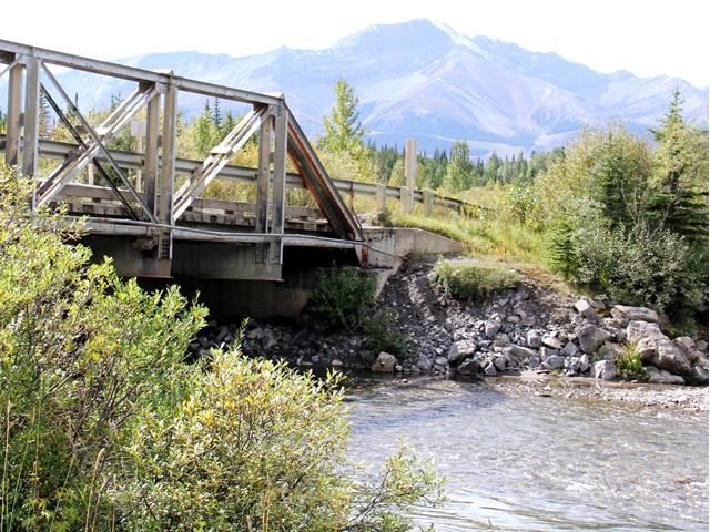Wooden bridge crossing a clear creek surrounded by greenery and mountains