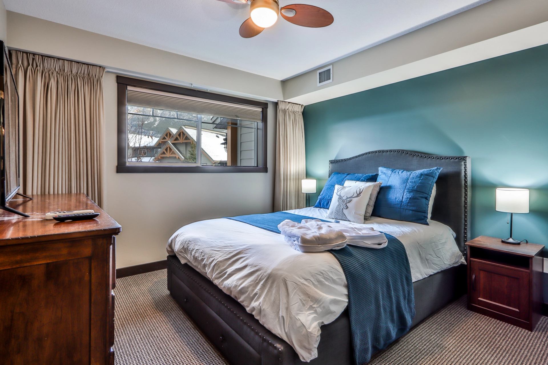 A bedroom with a bed featuring blue and white bedding, a teal accent wall, wooden dresser with a TV, and a window.