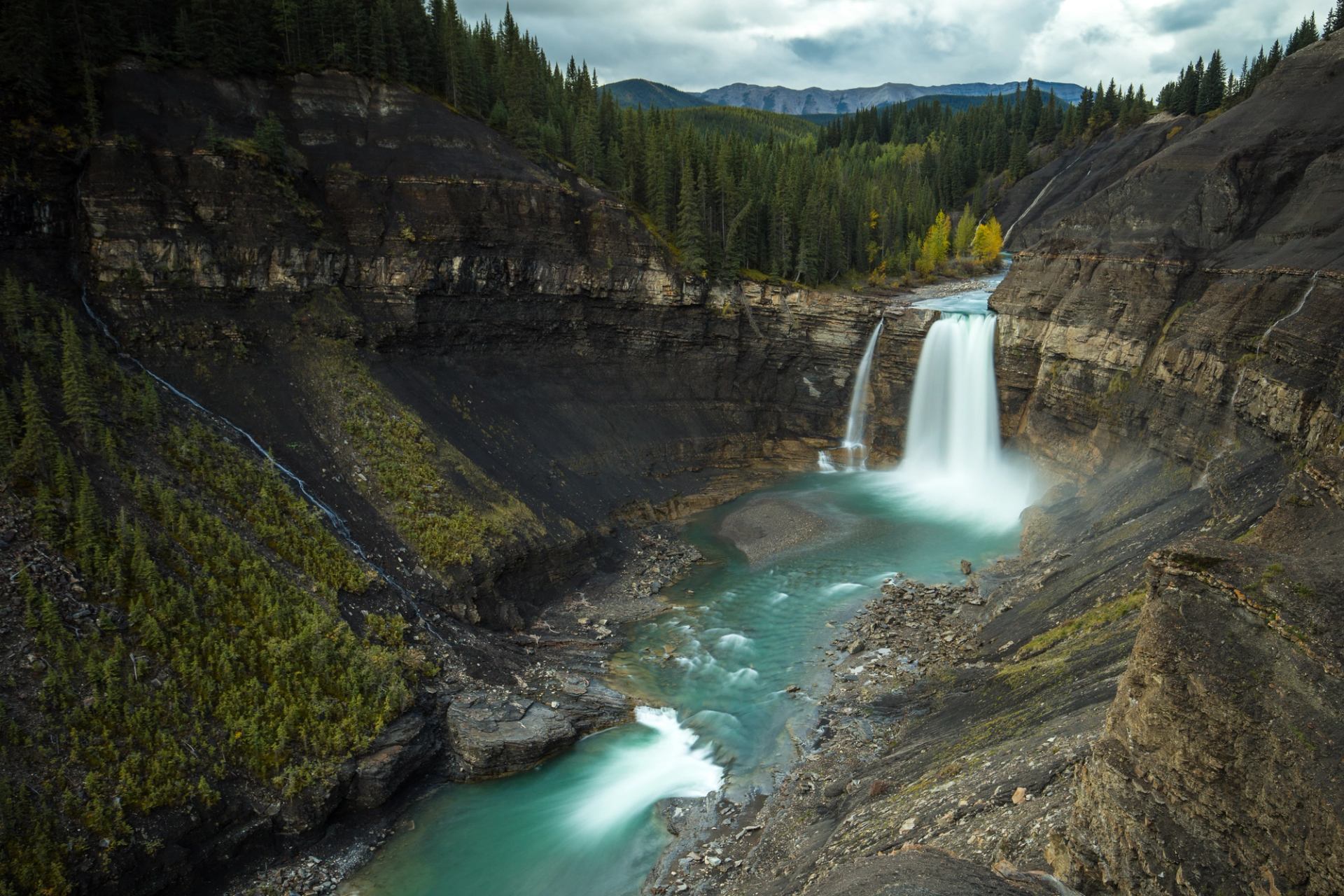 Turquoise river flows through rocky canyon with twin waterfalls and dense evergreen forest.