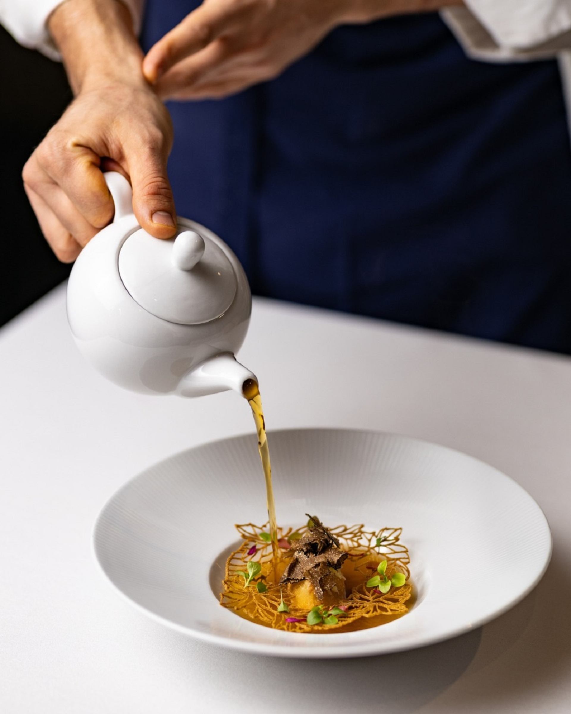 White teapot pours broth over plated gourmet dish with truffle and garnishes.