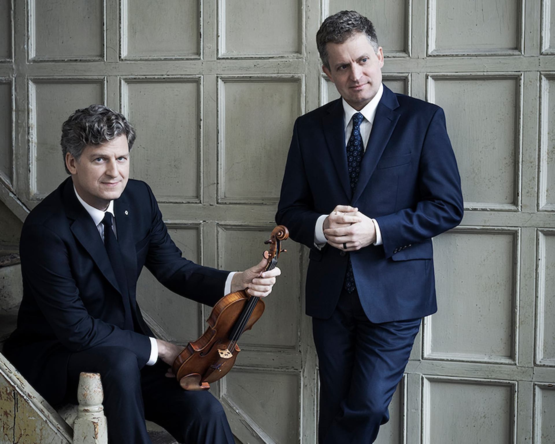 Two people in dark suits pose indoors, one seated holding a violin.