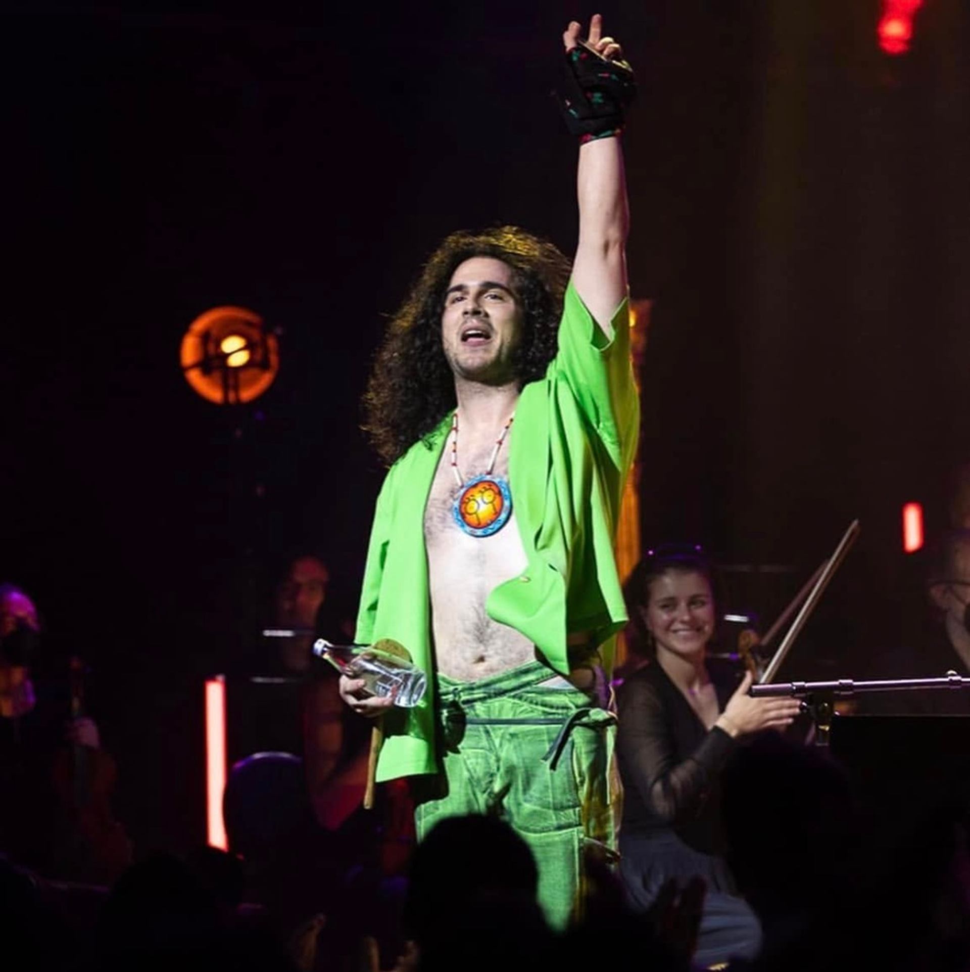 Performer in bright green raises one arm onstage beside a seated violinist.