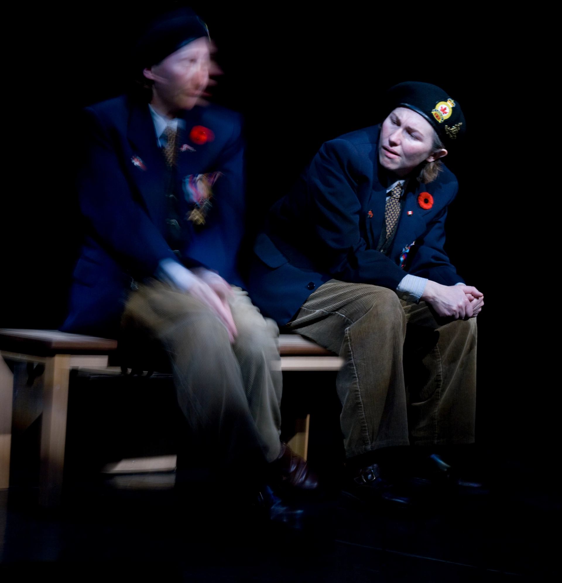 Two people in berets and blazers with poppies sit on a bench onstage.